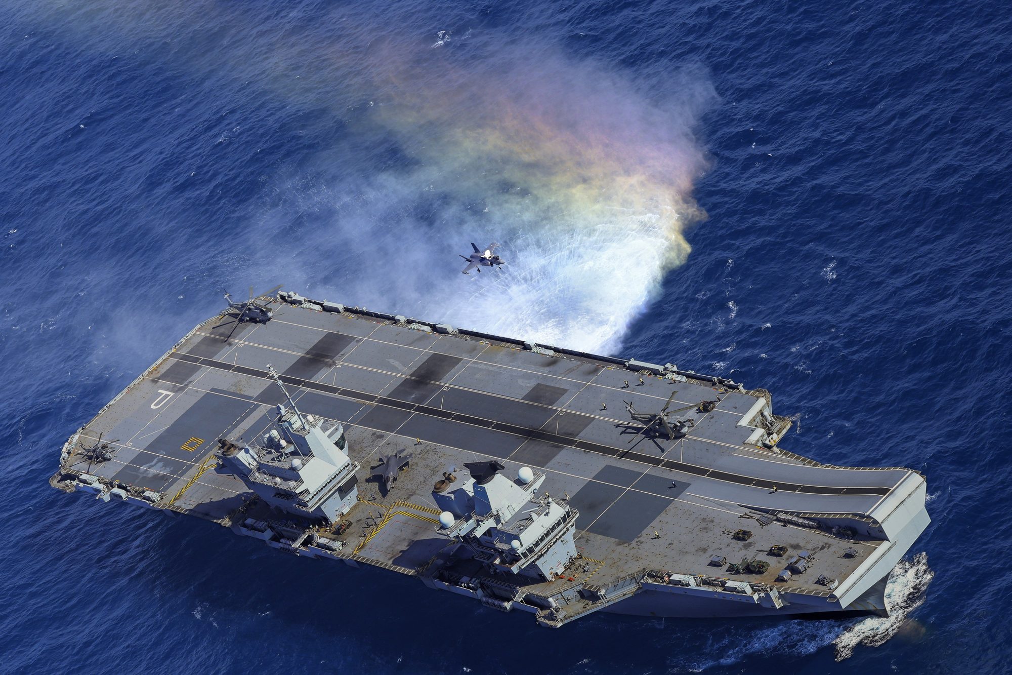 Aerial view of a Royal Navy aircraft carrier with an F-35 in flight alongside.
